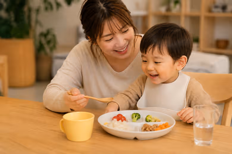 温かいキッチンの食卓で、保護者がスマートフォンを持ち、服を着た幼児と食事をする穏やかな日常